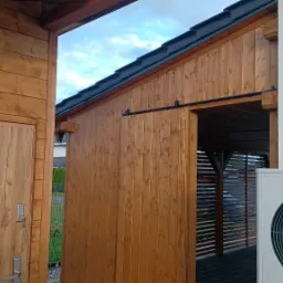 Drewniana konstrukcja z przesuwanymi drzwiami, widoczny fragment dachu pokrytego dachówką, obok biała ściana z rurą spustową i jednostką zewnętrzną pompy ciepła.