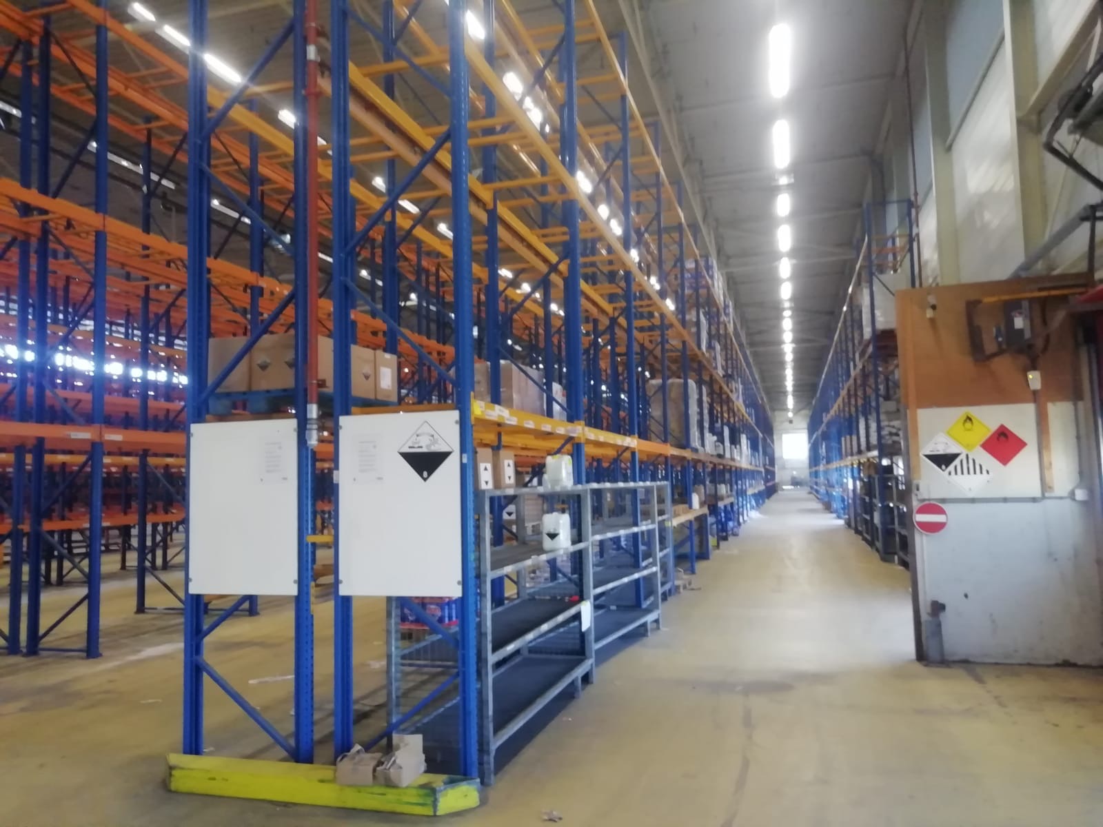 Wnętrze magazynu z wysokimi, metalowymi regałami w kolorach niebieskim i pomarańczowym, wypełnionymi kartonami i pojemnikami, z widocznymi tablicami ostrzegawczymi o substancjach niebezpiecznych.