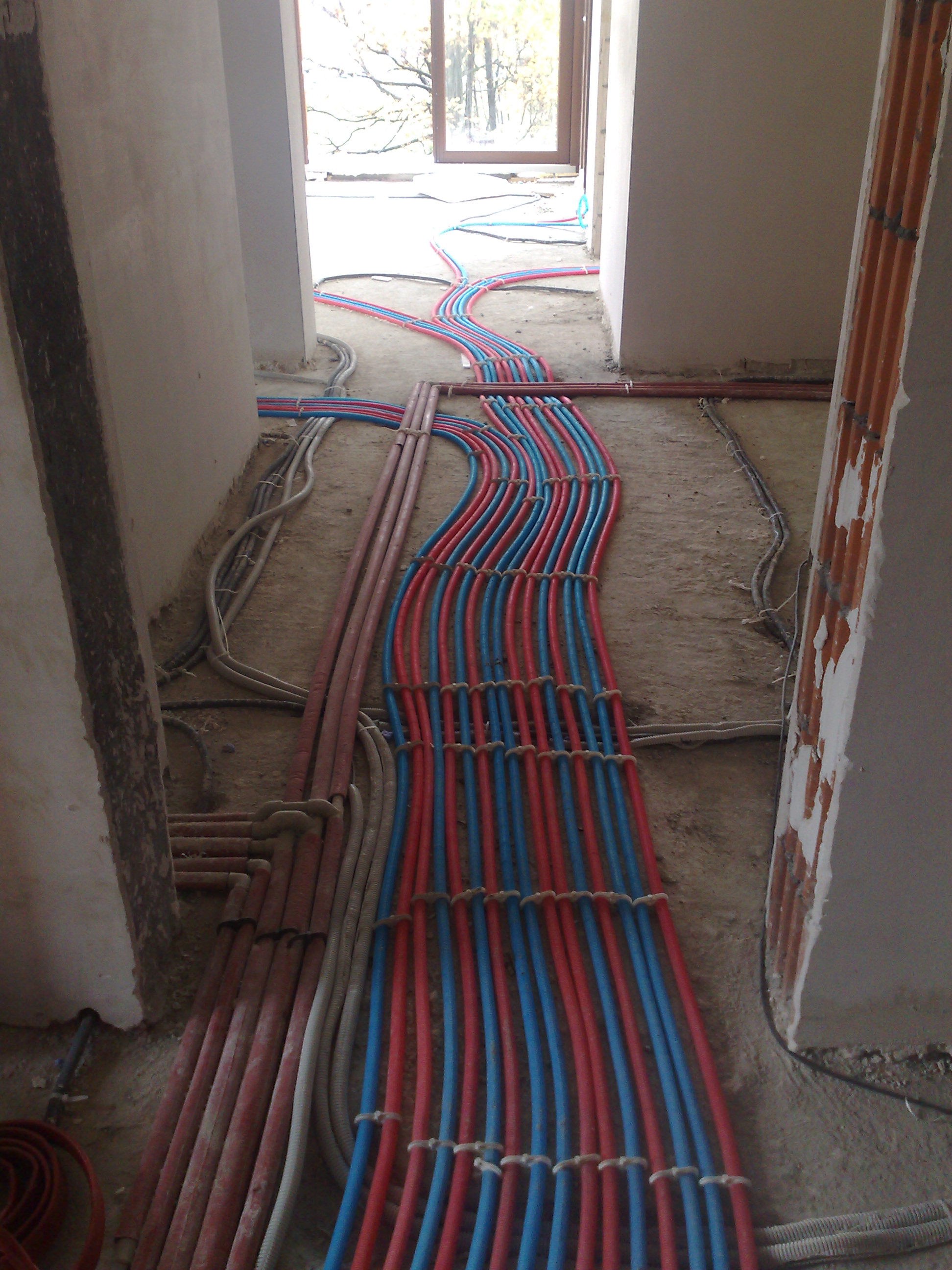 Równolegle ułożone, spięte w regularnych odstępach rury instalacyjne w kolorach czerwonym i niebieskim, prowadzone po surowej podłodze w trakcie budowy.