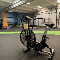 Airbike z wentylatorem w siłowni we Wrocławiu; w tle widoczne inne urządzenia do ćwiczeń, takie jak bieżnia, sztangi i drabinki.