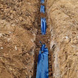 INSTALPRO ARTUR MIERZWIŃSKI - Wykopany rów w ziemi z ułożonym niebieskim kablem elektrycznym w osłonie, widoczna koparka Terex i budynek w trakcie budowy w tle.