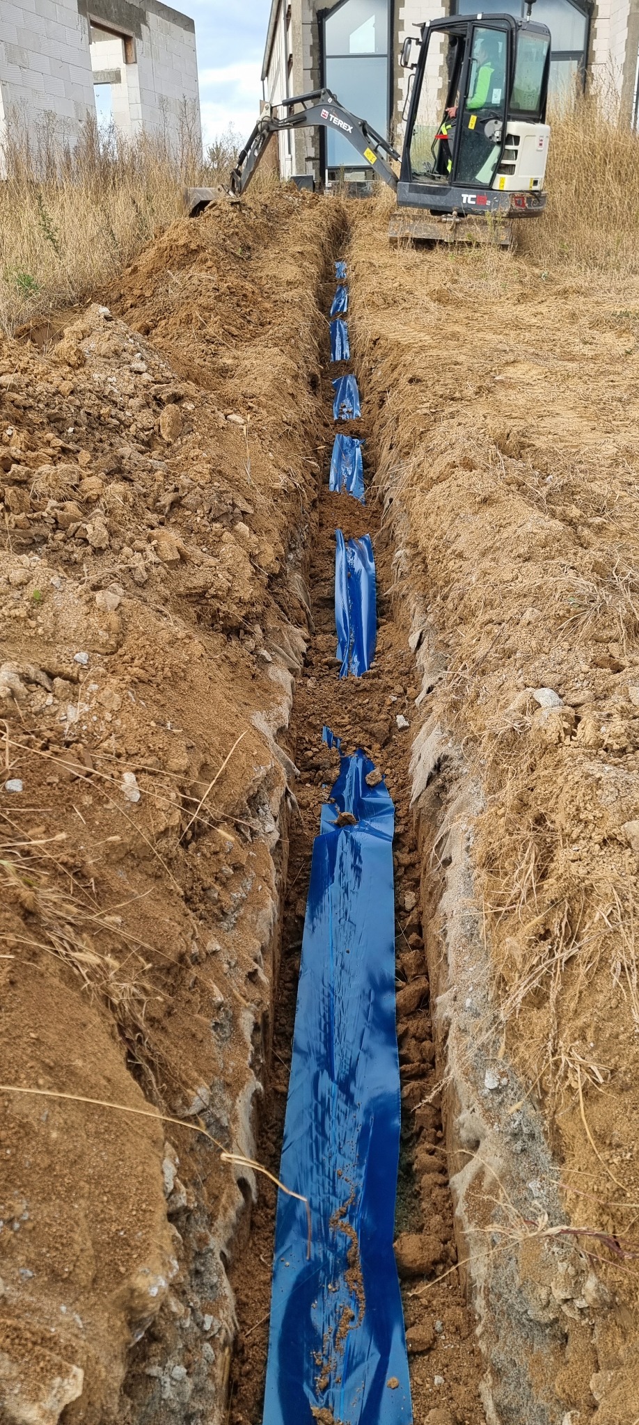 Wykopany rów w ziemi z ułożonym niebieskim kablem elektrycznym w osłonie, widoczna koparka Terex i budynek w trakcie budowy w tle.