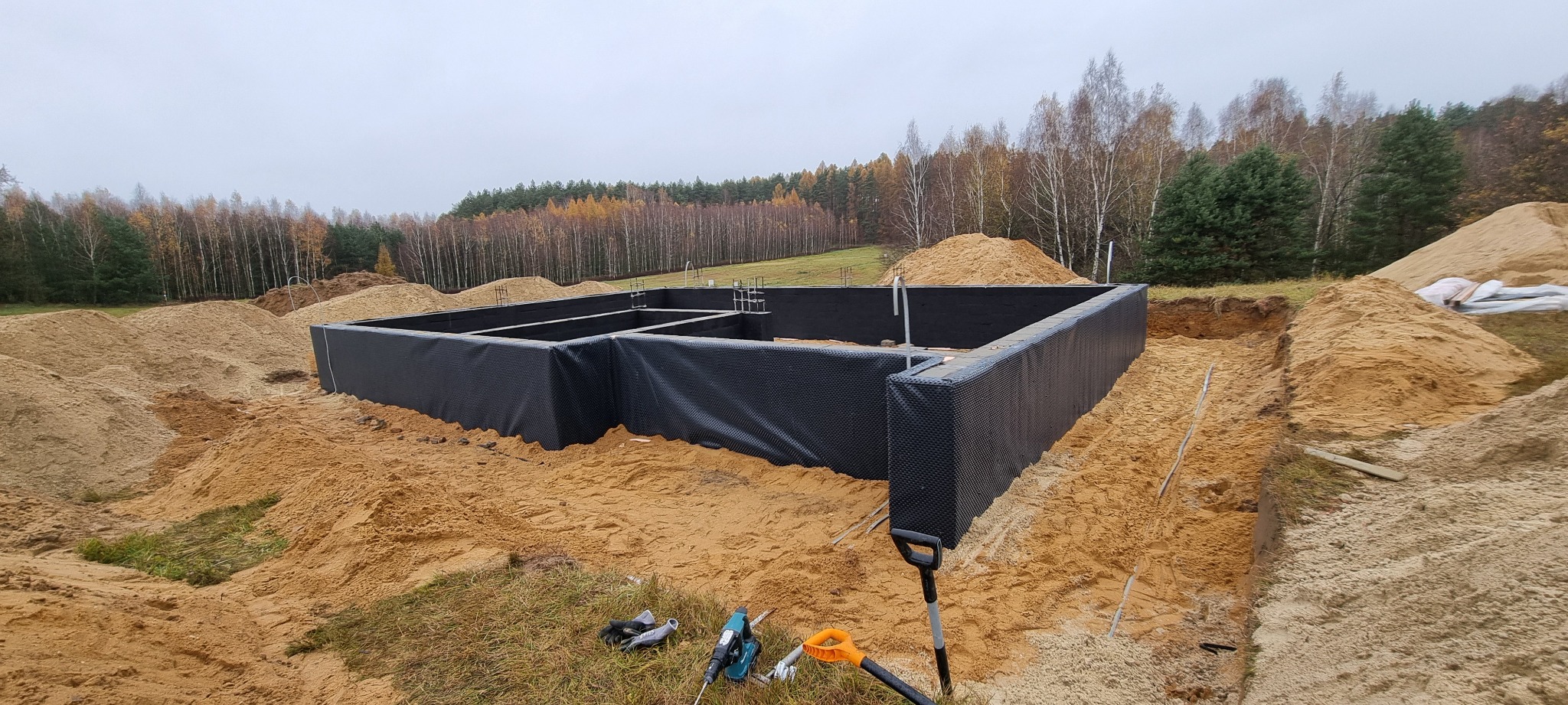 Fundamenty domu jednorodzinnego z izolacją przeciwwilgociową, widoczne narzędzia budowlane i hałdy piasku na działce.