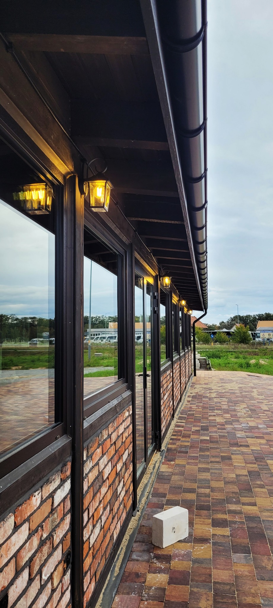 Zewnętrzne oświetlenie budynku z ceglaną elewacją i dużymi oknami, widoczne lampy w stylu retro zamontowane pod okapem, brązowe rynny i kostka brukowa na chodniku.