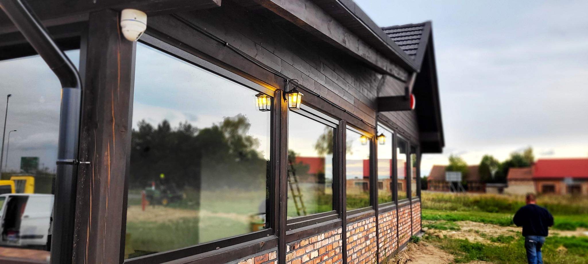 Zewnętrzny widok drewnianej altany z oknami i oświetleniem w postaci stylizowanych latarenek, z widocznym fragmentem instalacji elektrycznej doprowadzającej prąd do lamp. Na zewnątrz widać...