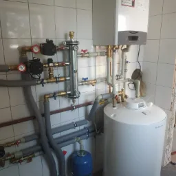 Kotłownia z zamontowanym piecem gazowym Ariston, zbiornikiem na wodę i osprzętem hydraulicznym na tle białych, kwadratowych płytek ściennych.