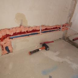 Rot Therm Instalatorstwo sanitarne - Instalacja hydrauliczna w ścianie z czerwonymi i niebieskimi rurami, zabezpieczona różową pianką montażową, widoczne narzędzia - młotek i rurki, surowy stan wykończenia.