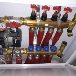 Rozdzielacz instalacji centralnego ogrzewania w białej szafce, widoczne czerwone rury, złote zawory z czerwonymi i niebieskimi pokrętłami, manometr i pompa obiegowa.