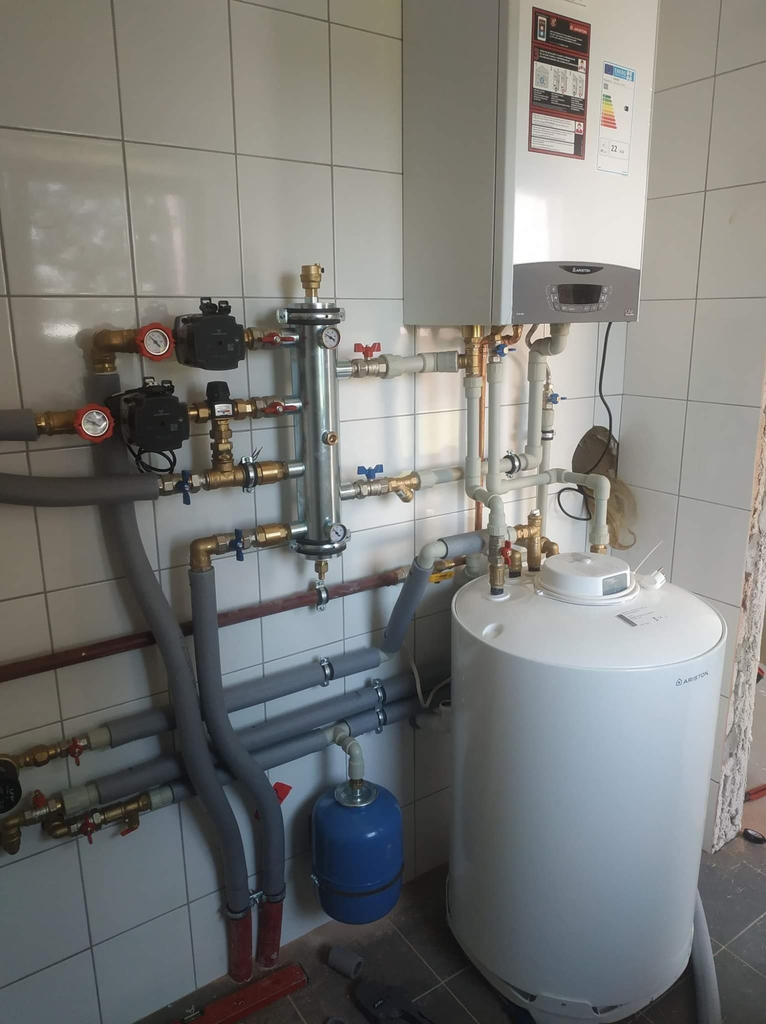 Kotłownia z zamontowanym piecem gazowym Ariston, zbiornikiem na wodę i osprzętem hydraulicznym na tle białych, kwadratowych płytek ściennych.