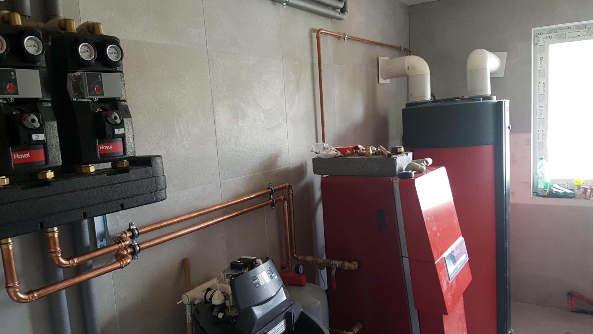 Częściowo zmontowana instalacja grzewcza z kotłem i rozdzielaczami Hoval, rurami miedzianymi oraz przyłączeniami, widoczne narzędzia na kotle i okno w tle.