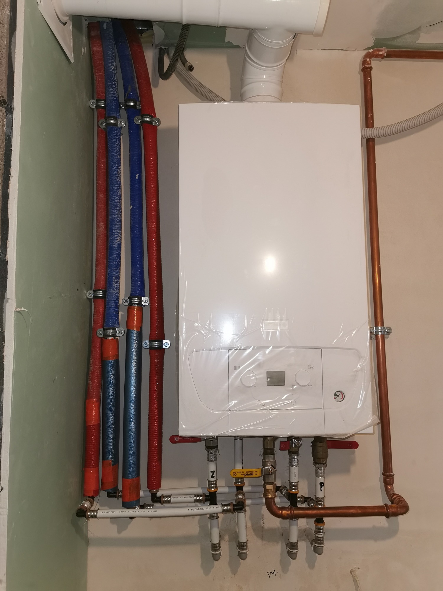 Nowy, biały piec centralnego ogrzewania, jeszcze w folii ochronnej, z podłączonymi rurami miedzianymi i plastikowymi, zamocowanymi do ściany za pomocą metalowych obejm.
