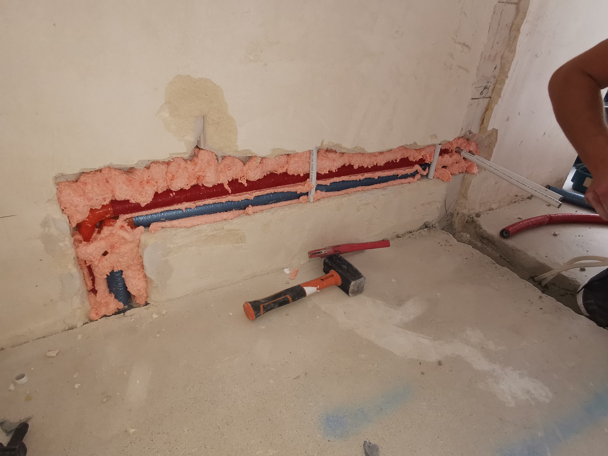Instalacja hydrauliczna w ścianie z czerwonymi i niebieskimi rurami, zabezpieczona różową pianką montażową, widoczne narzędzia - młotek i rurki, surowy stan wykończenia.