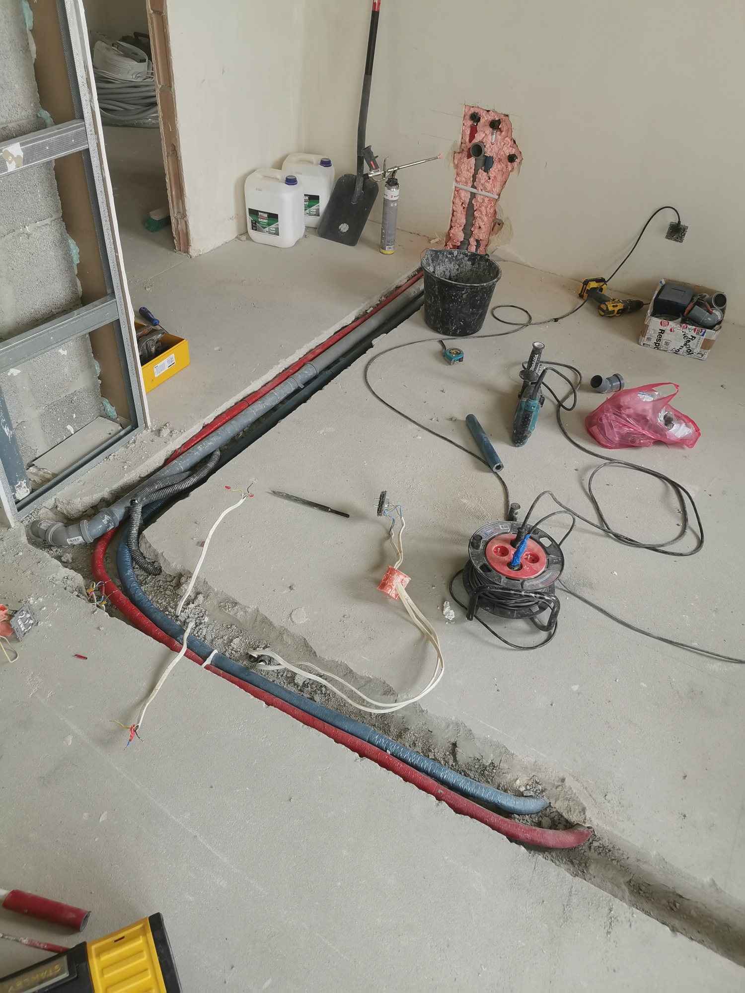 Wykop w posadzce z ułożonymi rurami instalacji wodnej i grzewczej, widoczne narzędzia i materiały budowlane w surowym wnętrzu.