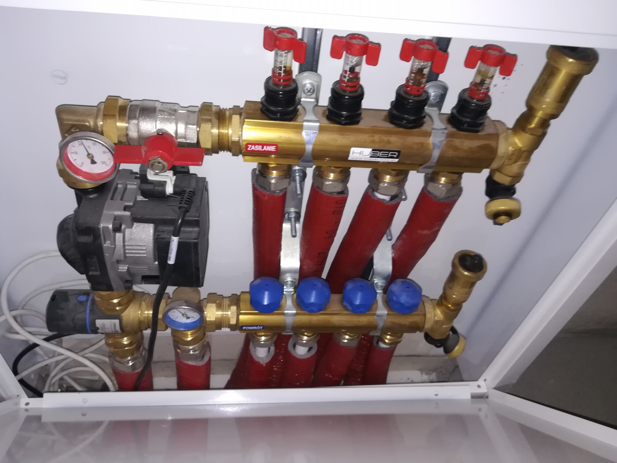 Rozdzielacz instalacji centralnego ogrzewania w białej szafce, widoczne czerwone rury, złote zawory z czerwonymi i niebieskimi pokrętłami, manometr i pompa obiegowa.