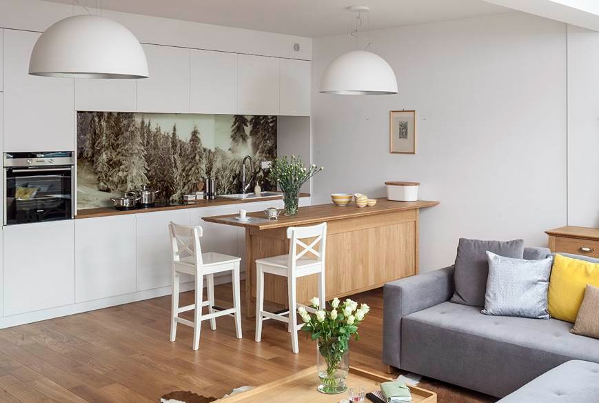 Jasne wnętrze: kuchnia z białymi szafkami i drewnianym blatem, krzesła barowe, sofa. Na ścianie fototapeta z motywem zimowego lasu. Nowoczesny design.