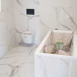 Max-Bud - Nowoczesna łazienka w trakcie prac wykończeniowych: białe płytki imitujące marmur na ścianach i podłodze, podwieszana toaleta z czarnym przyciskiem spłukującym, zabudowana wanna częściowo...