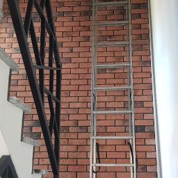 Max-Bud - Ściana wyłożona czerwoną cegłą klinkierową z widoczną instalacją elektryczną, oparta o nią aluminiowa drabina, fragment schodów z czarną balustradą i karton na marmurowym podeście.