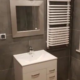 Umywalka z szafką w białym kolorze, lustro w srebrnej ramie, grzejnik drabinkowy i kryształowy kinkiet na szarej ścianie z płytek w łazience po remoncie.