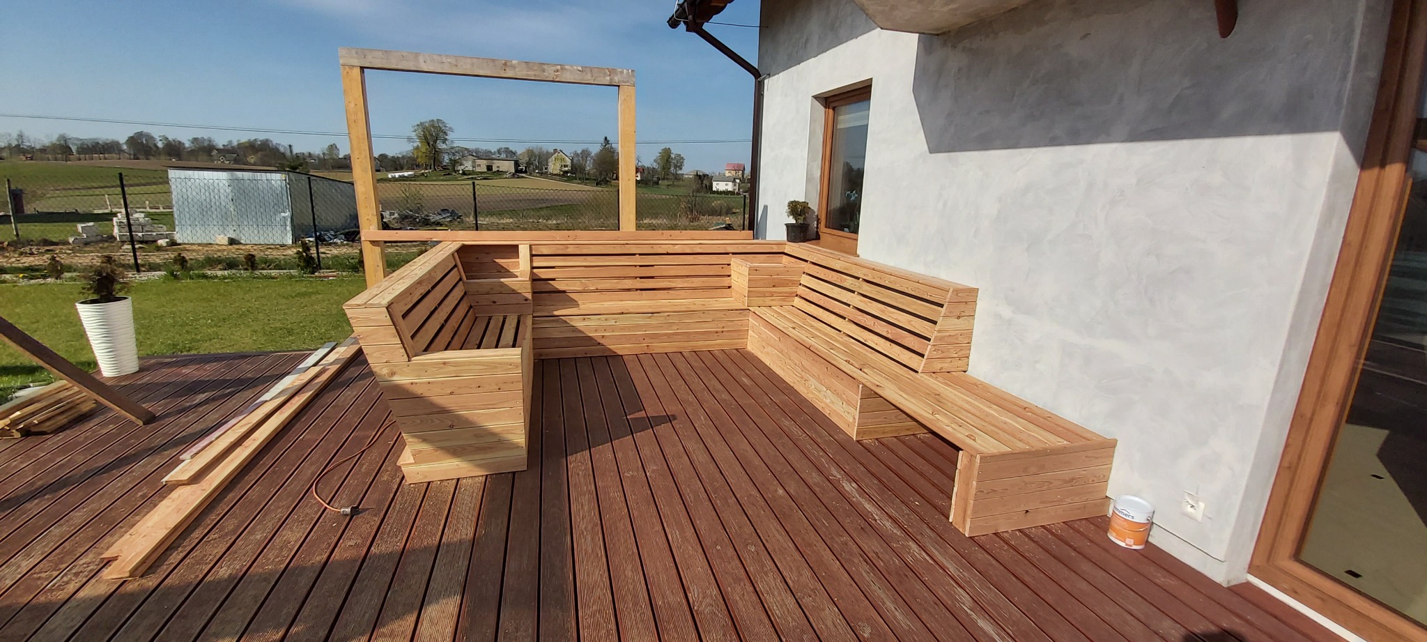 Nowoczesny taras z drewnianą podłogą i narożną zabudową siedziska z surowego drewna, widok na wiejski krajobraz w tle, elementy konstrukcyjne pergoli w trakcie montażu.