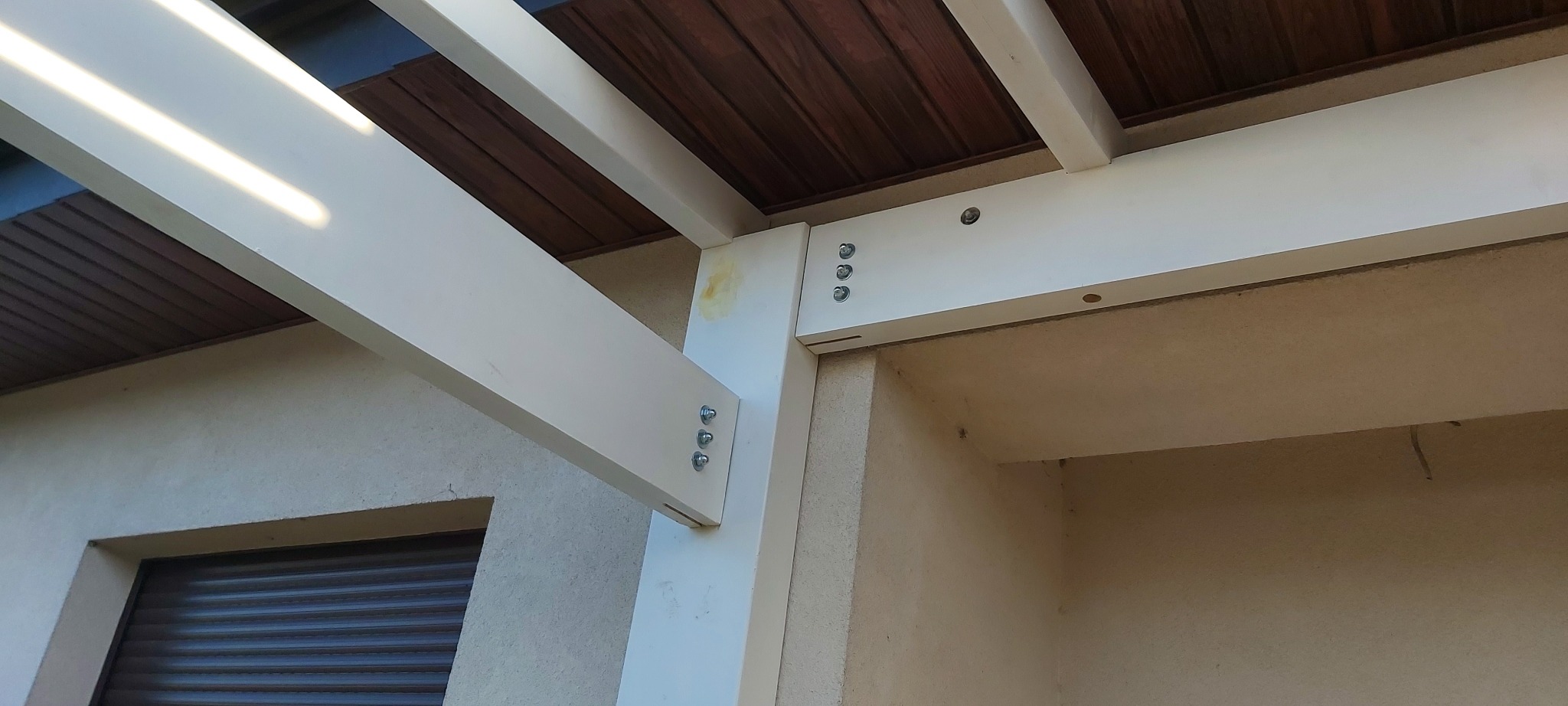 Fragment konstrukcji drewnianej, prawdopodobnie zadaszenia, z widocznymi połączeniami śrubowymi i bejcowaną drewnianą podsufitką.