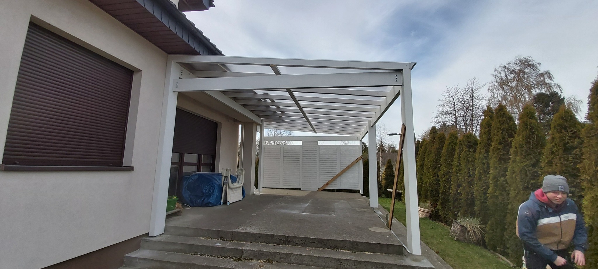 Nowoczesna, biała pergola z przezroczystym zadaszeniem, przylegająca do domu w Załęże, z widocznymi elementami konstrukcyjnymi i tłem z drzew i krzewów.