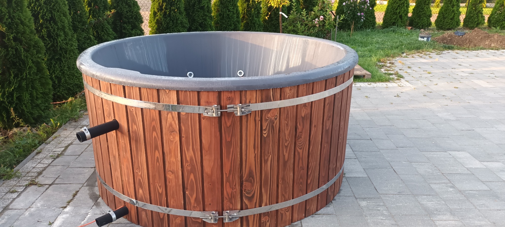 Drewniana balia ogrodowa z szarą wkładką, wzmocniona metalowymi obręczami, na tle kostki brukowej i zieleni.
