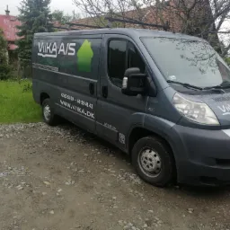Szary bus firmy VIKA A/S z logo i numerem telefonu zaparkowany na żwirowej drodze przed domem.