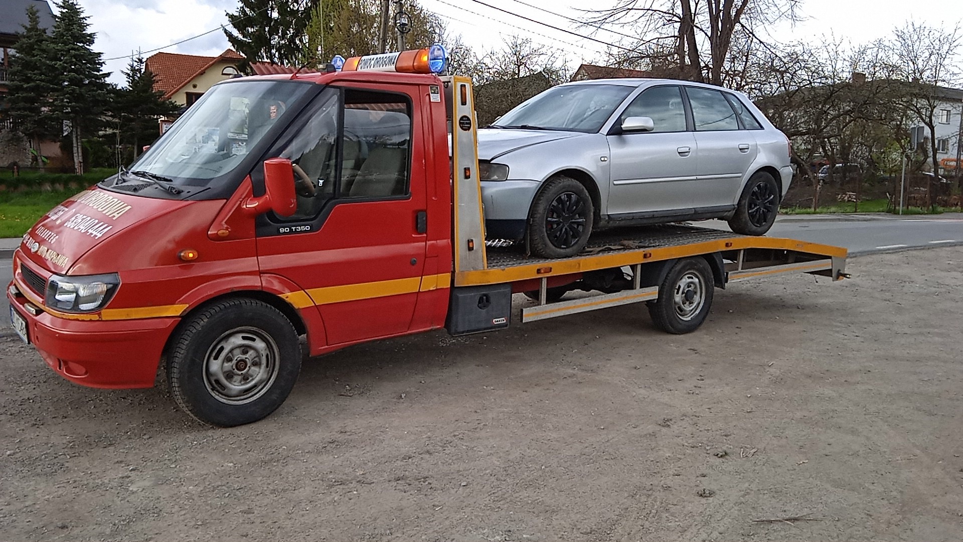 Czerwony Ford Transit T350 z lawetą przewozi srebrny samochód osobowy na tle zabudowy jednorodzinnej.