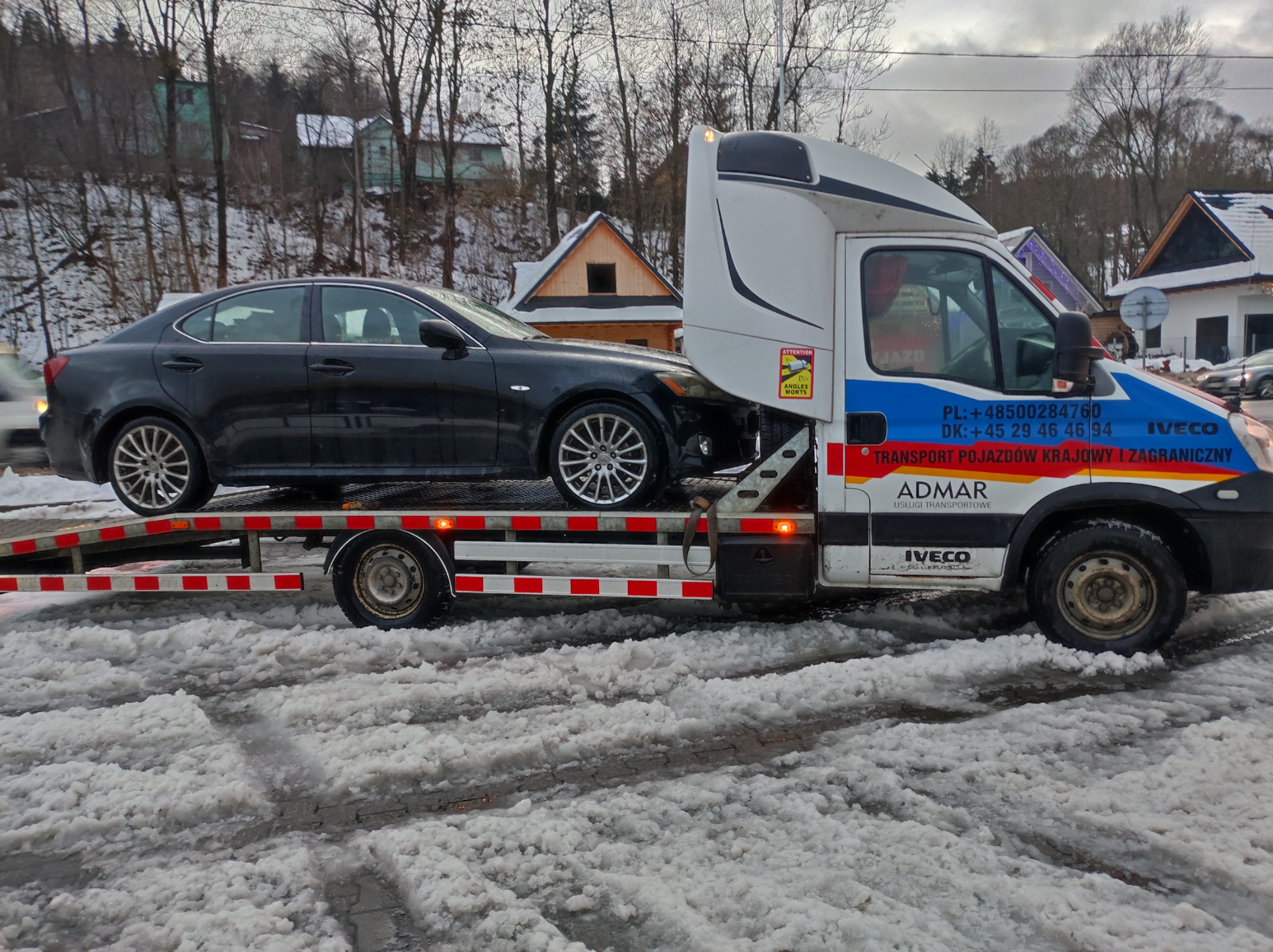 Czarny samochód osobowy marki Lexus przewożony na lawecie w zimowych warunkach; widoczne budynki w tle