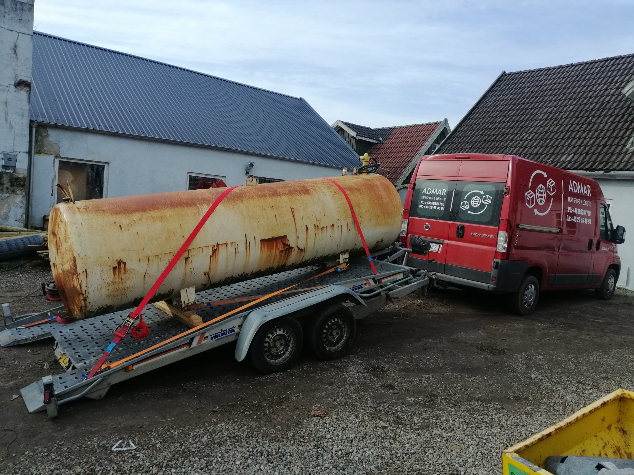Cylindryczny, zardzewiały zbiornik zabezpieczony czerwonymi pasami na dwuosiowej przyczepie, ciągniętej przez czerwonego busa firmy transportowej ADMAR na tle budynków.