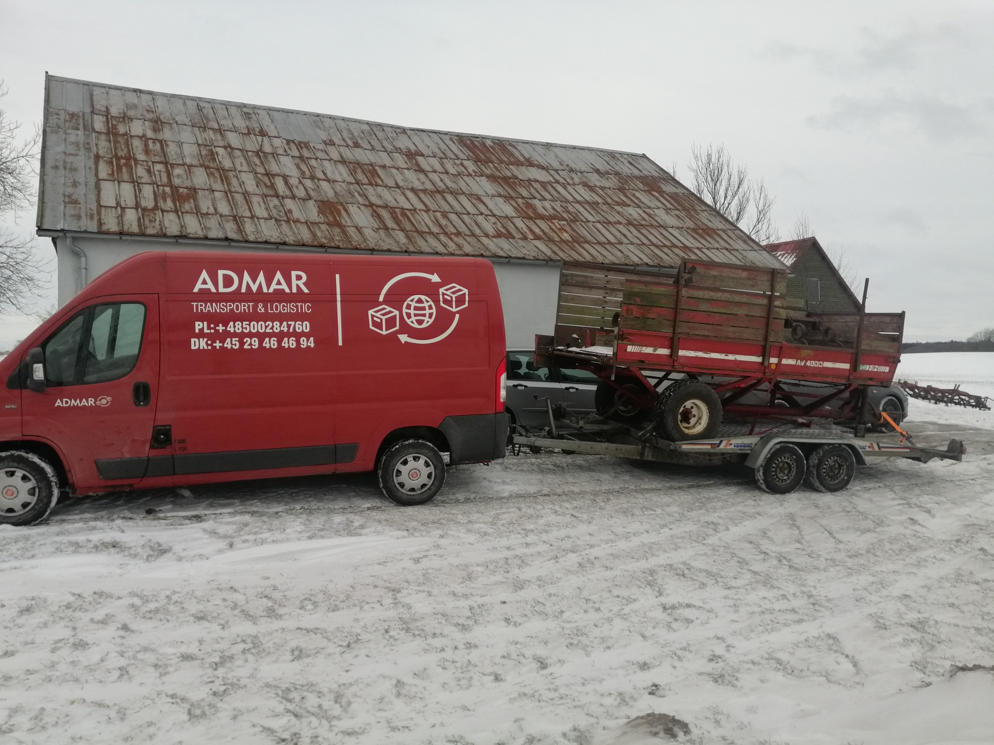Czerwony bus firmy ADMAR z przyczepą przewożącą platformę rolniczą na tle zaśnieżonego pola i budynku gospodarczego.