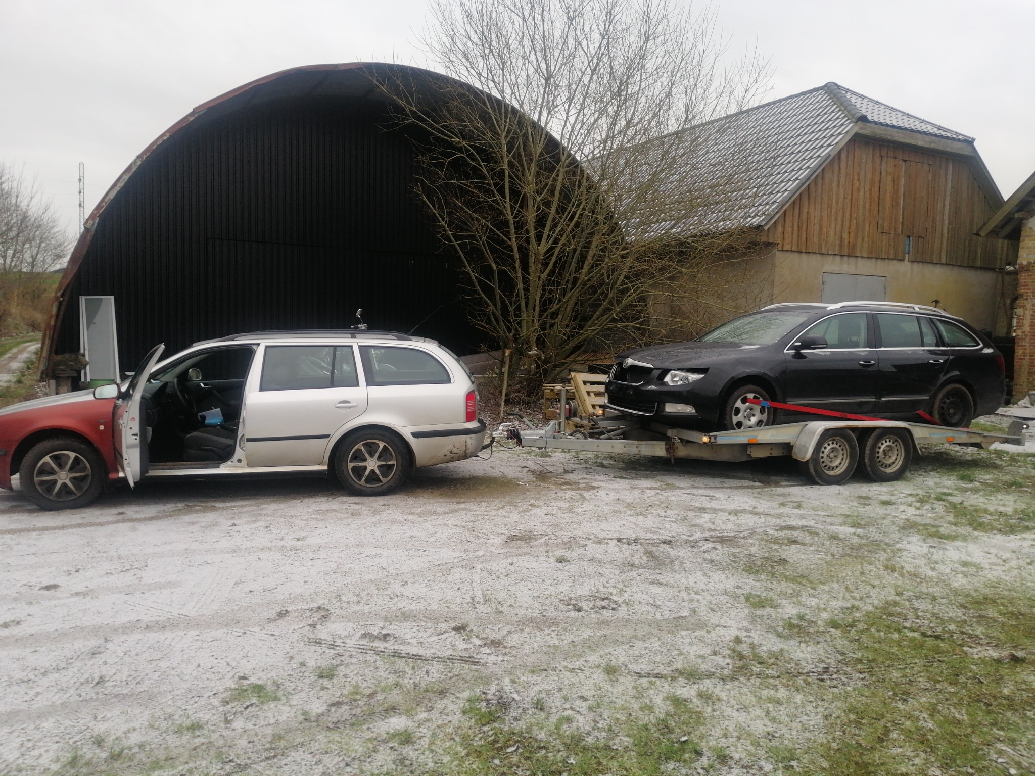Srebrny samochód osobowy z otwartymi drzwiami holuje na przyczepie lawetowej czarny samochód osobowy na tle starego budynku gospodarczego.