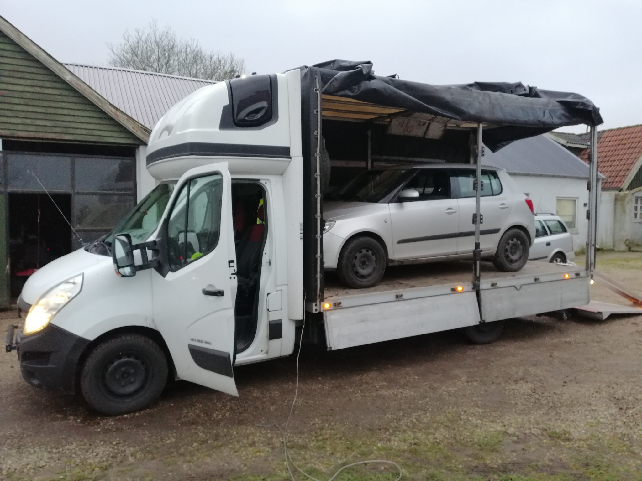 Biały samochód dostawczy z otwartymi drzwiami, na platformie załadowany srebrny samochód osobowy, tło wiejskie zabudowania.