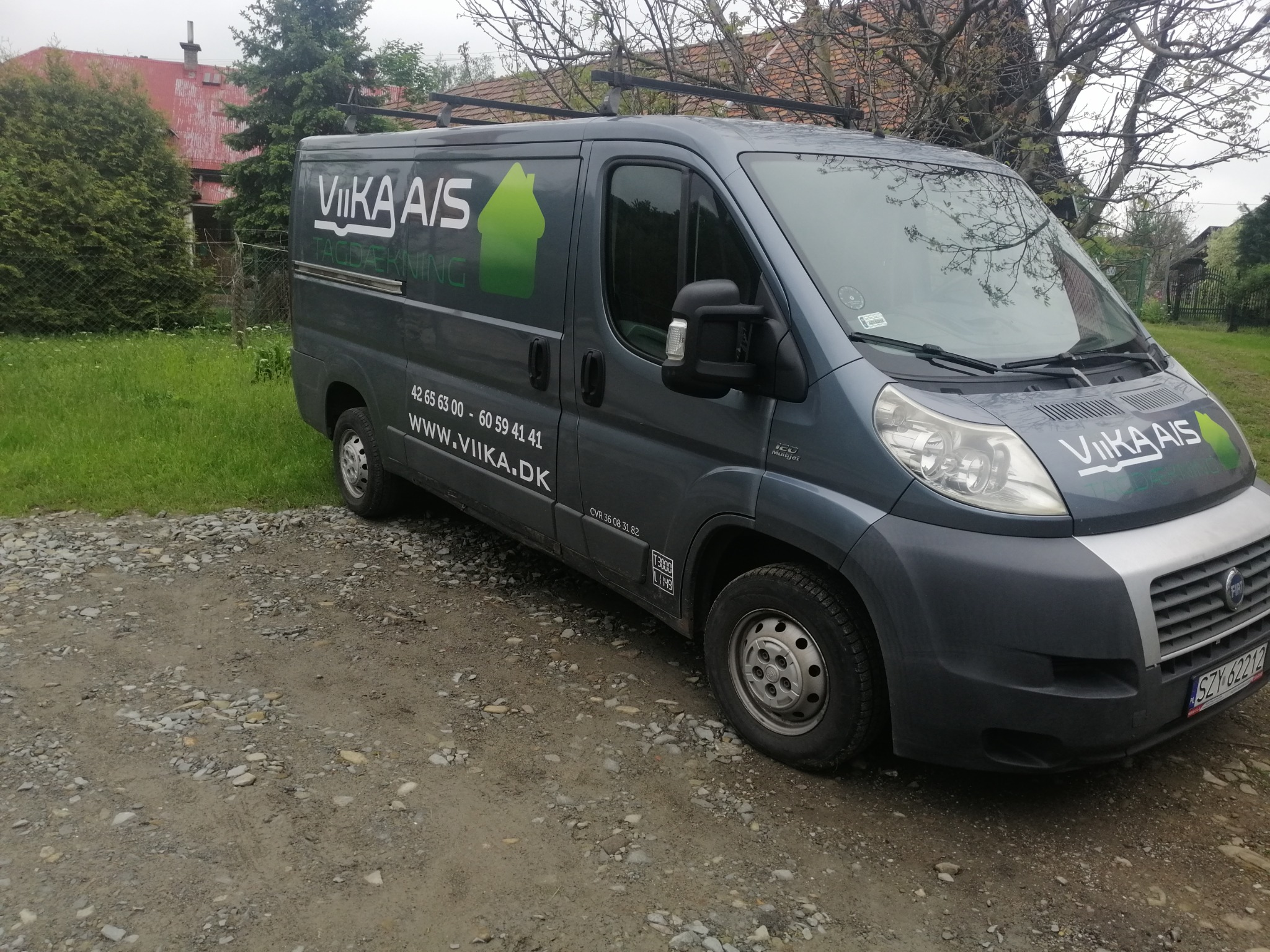Szary bus firmy VIKA A/S z logo i numerem telefonu zaparkowany na żwirowej drodze przed domem.
