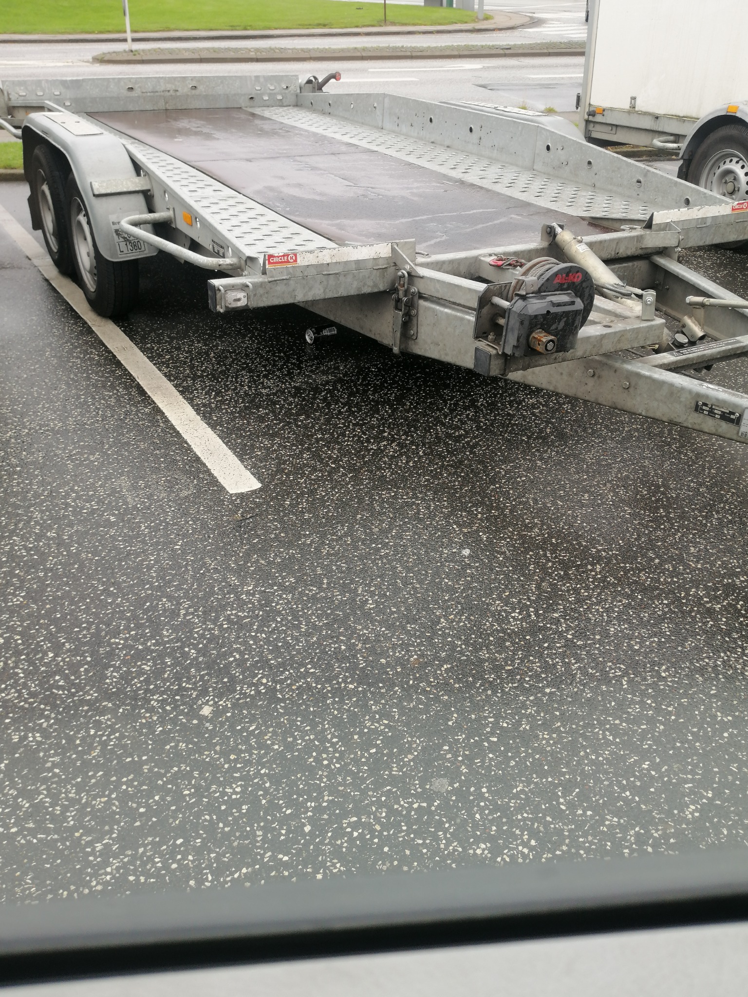 Szara, pusta laweta samochodowa z wyciągarką, zaparkowana na mokrym parkingu w pochmurny dzień.