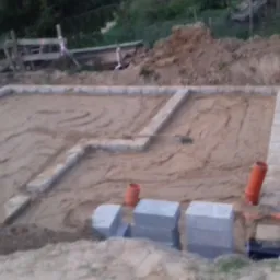 Przygotowany teren pod wylewkę fundamentów z wytyczonym obrysem budynku, widoczne cegły klinkierowe, rury PCV oraz szpadel z pomarańczowym uchwytem.