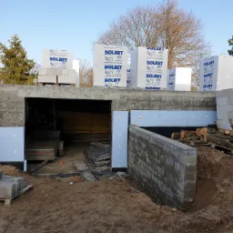 Budowa domu w trakcie: wzniesione ściany z bloczków betonowych, częściowo ocieplone, palety z materiałami budowlanymi Solbet na stropie, widoczny wykop ziemny i drewno opałowe.