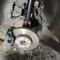 Zdjęcie tarczy hamulcowej z zaciskiem i elementami zawieszenia w samochodzie podczas naprawy, widoczne ślady użytkowania i zabrudzenia.