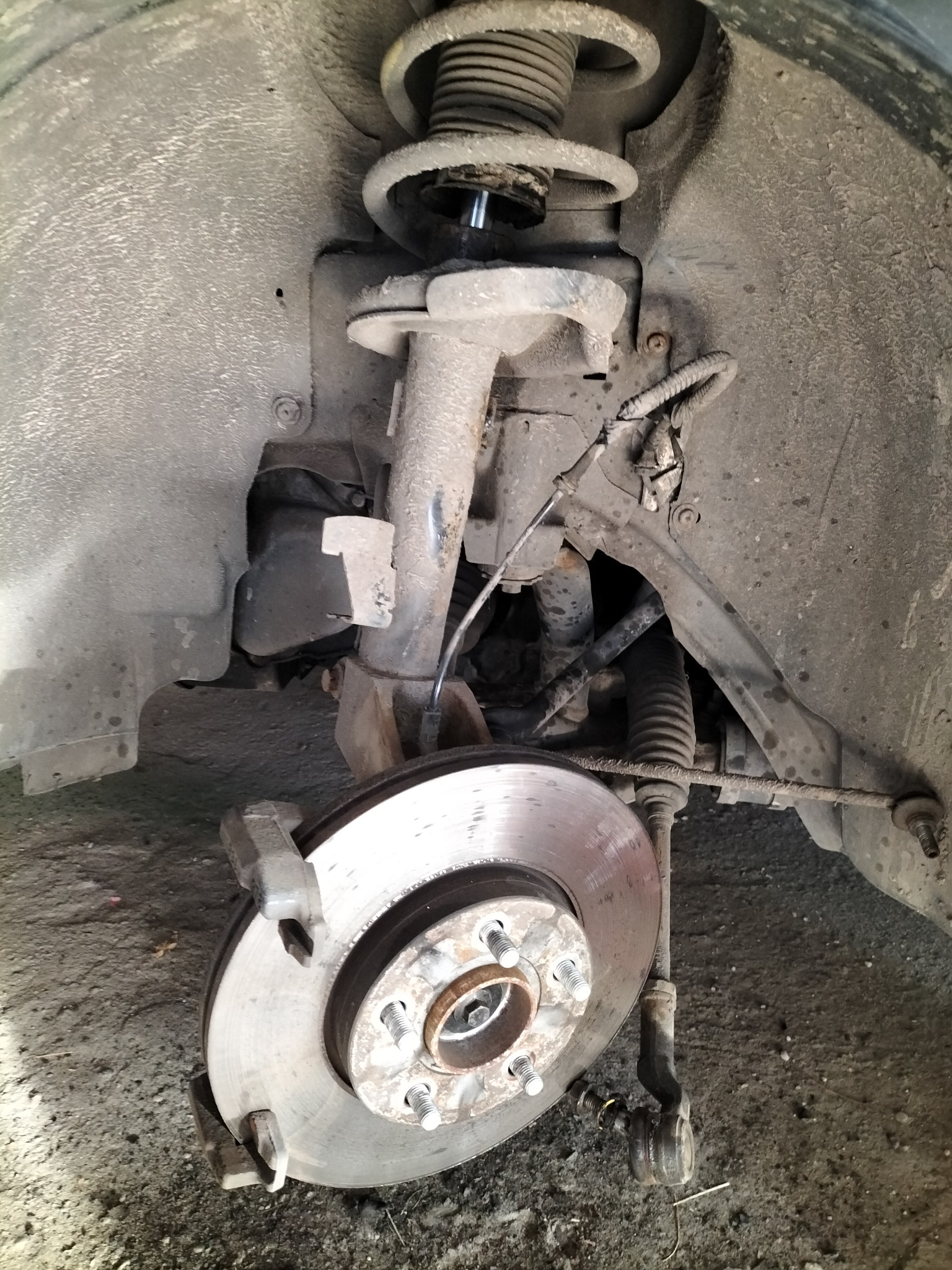 Brudne zawieszenie samochodowe z tarczą hamulcową i sprężyną amortyzatora w trakcie naprawy, widoczne elementy układu kierowniczego i osłona nadkola.