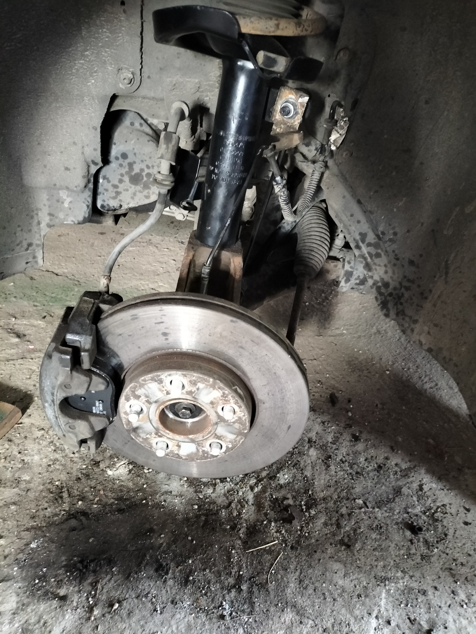 Zdjęcie tarczy hamulcowej z zaciskiem i elementami zawieszenia w samochodzie podczas naprawy, widoczne ślady użytkowania i zabrudzenia.