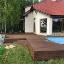 Drewniany taras przylegający do domu z basenem, obok zielony trawnik z konewką, w tle brzozowy lasek.