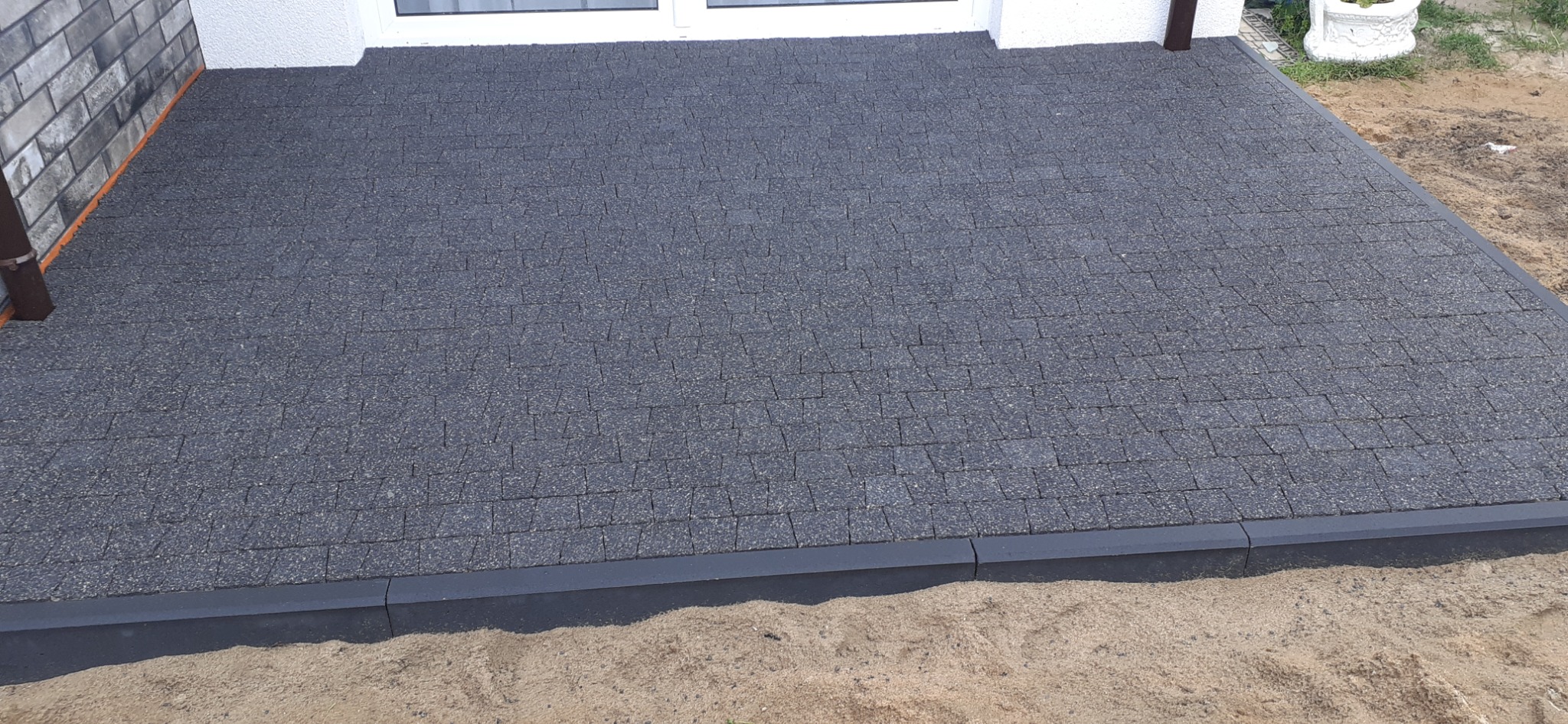 Ciemnoszara kostka brukowa ułożona na tarasie przed budynkiem, obrzeża wykończone ciemną kostką krawężnikową, widoczny fragment ściany i rynny.