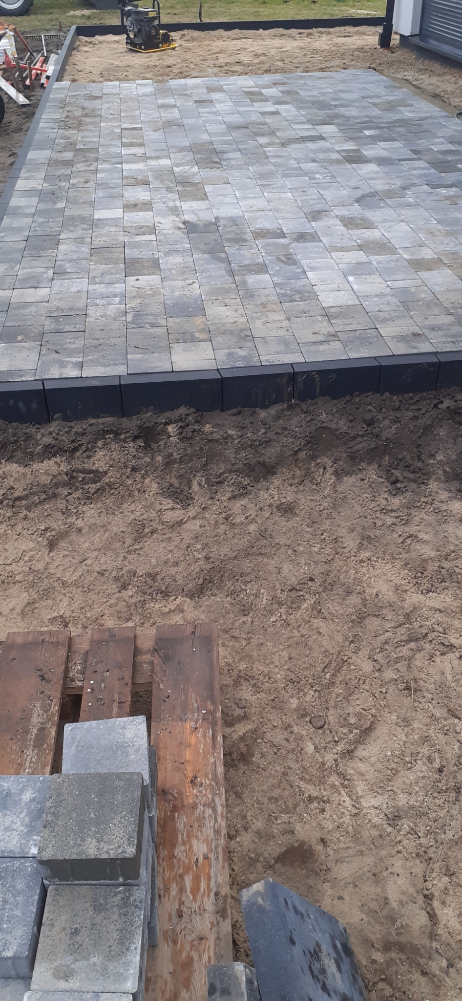 Wyrównana powierzchnia z szarej kostki brukowej z czarnym obramowaniem, widoczny fragment palety z kostką i ubijarka w tle.