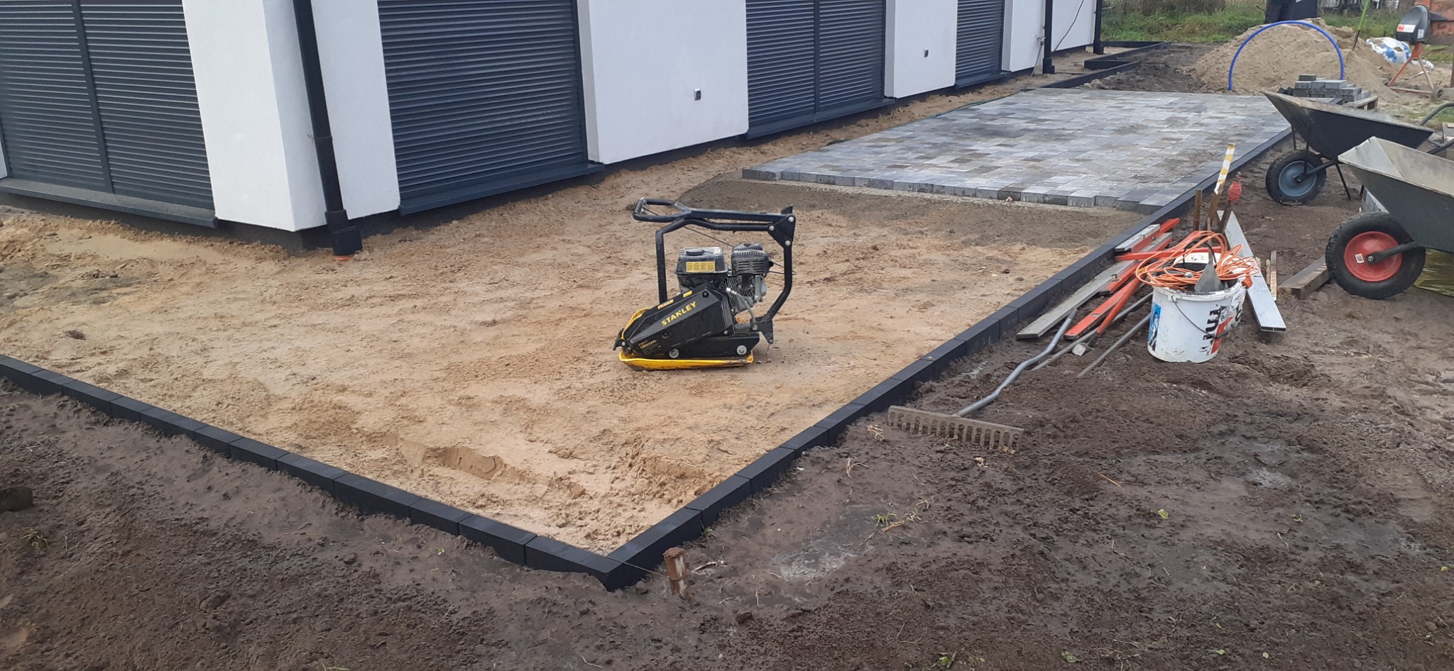 Żółto-czarna zagęszczarka Stanley na piasku, obrzeża z kostki, częściowo ułożona kostka brukowa w tle, przygotowanie terenu pod kostkę brukową.