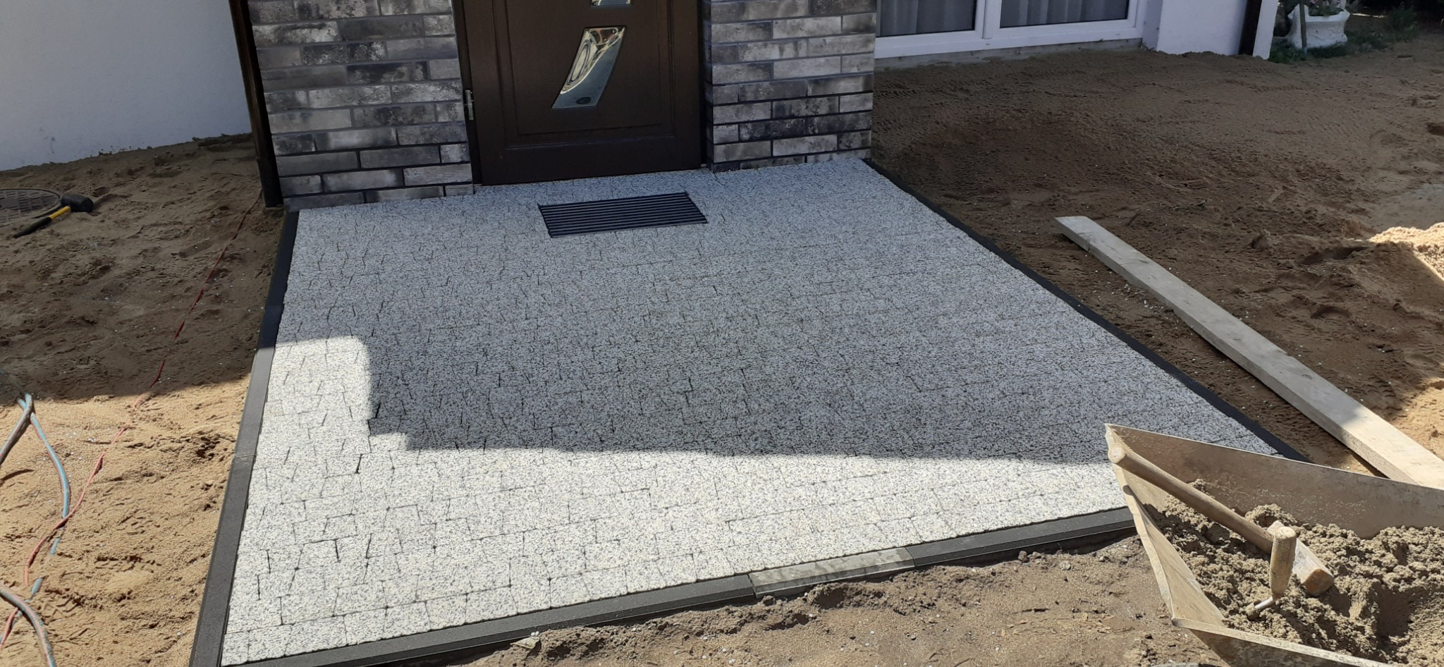Nowo ułożona, jasnoszara kostka przed wejściem do domu z ciemną, metalową kratką odpływową i fragmentem drzwi wejściowych; widoczny mur z szarej cegły, narzędzia budowlane i piasek.