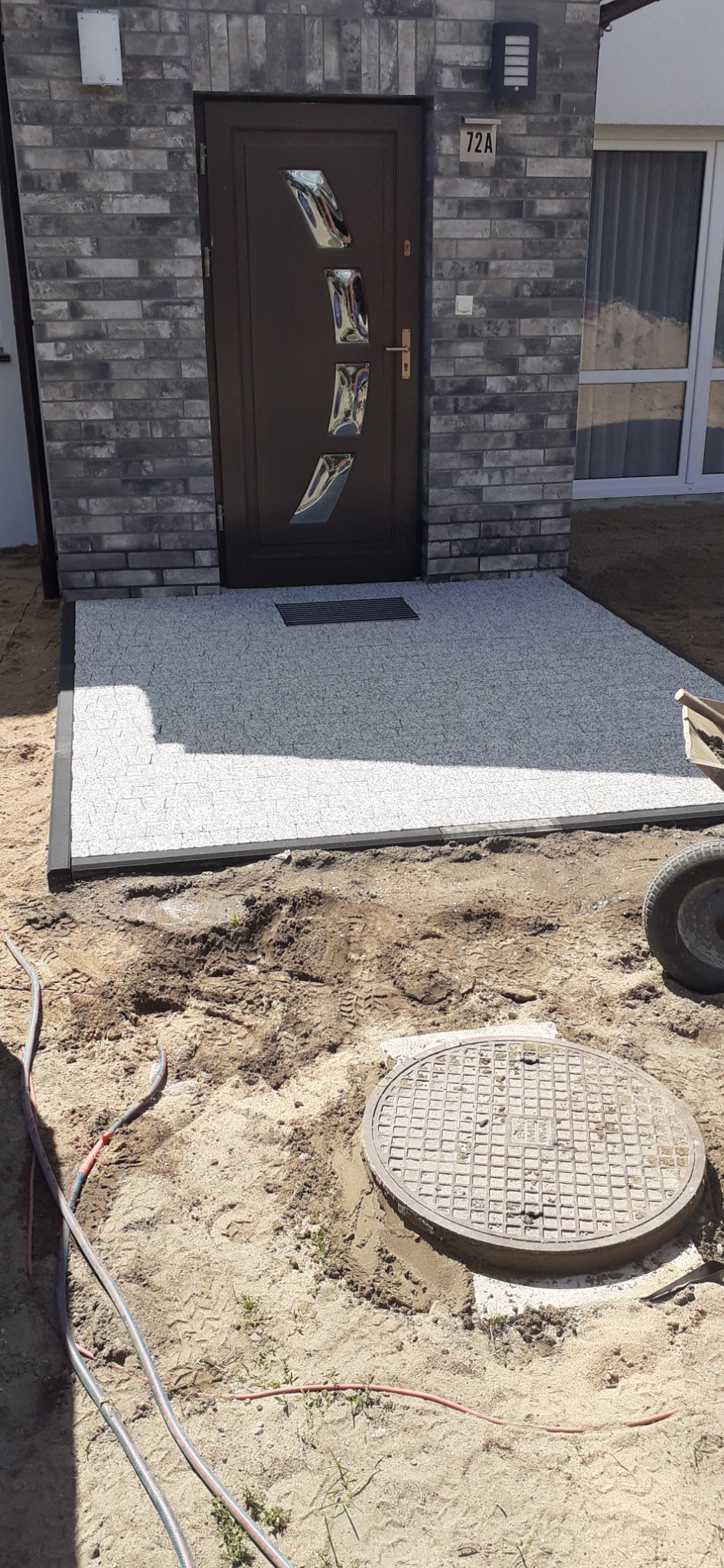 Wejście do domu z nową, jasną nawierzchnią z drobnych kamyczków, obrzeża w kolorze antracytowym, w tle elewacja z szarej cegły i brązowe drzwi z metalowymi zdobieniami, widoczny właz kanalizacyjny...