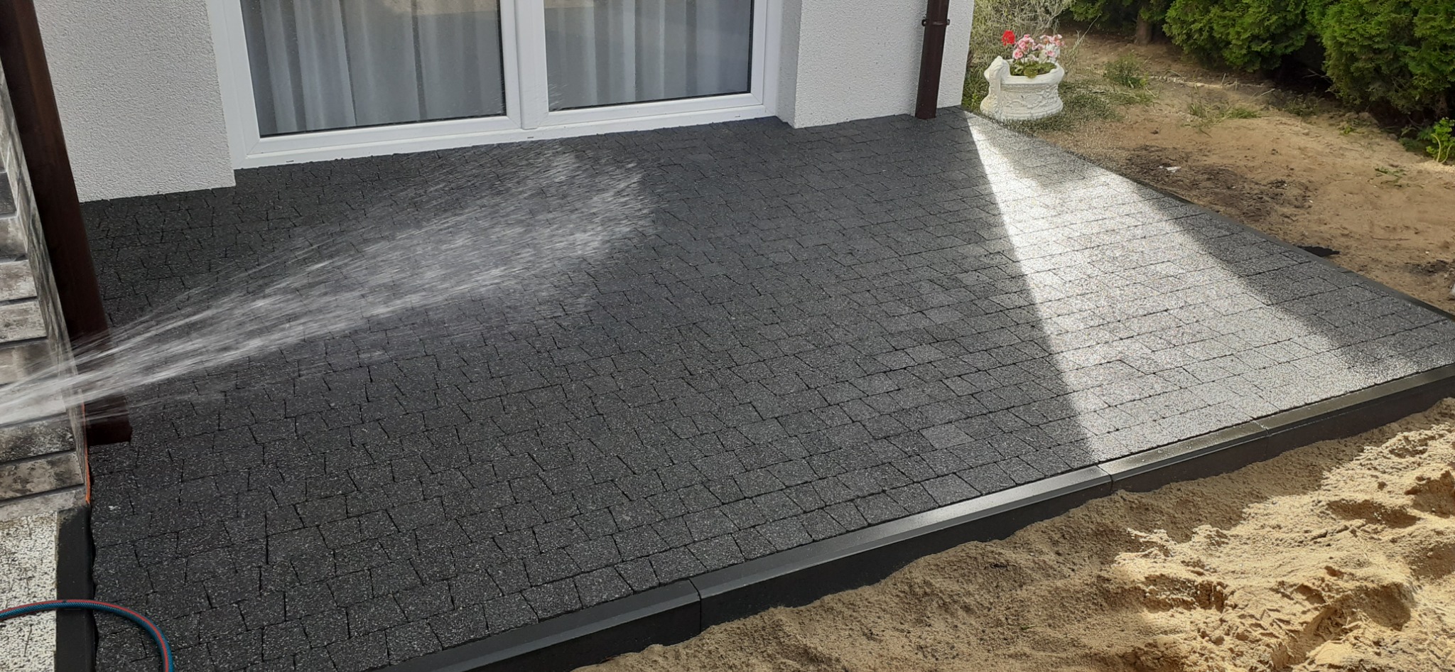 Czyszczenie ciśnieniowe nowo ułożonej, szarej kostki brukowej na posesji, widoczny wąż ogrodowy i strumień wody.