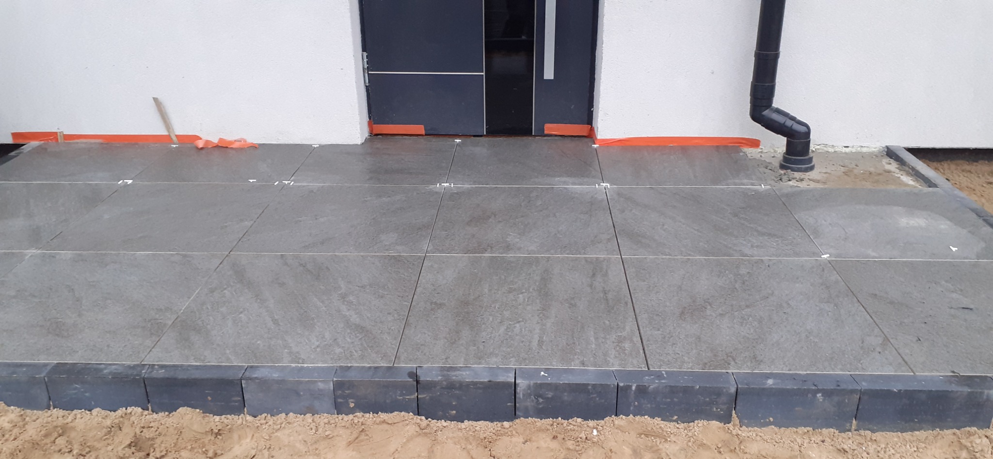 Ułożona szara płytka gresowa na zewnątrz budynku, z widocznymi fugami i obrzeżem z ciemnej kostki, zabezpieczona folią malarską przy ścianie i drzwiach wejściowych, w trakcie prac wykończeniowych.