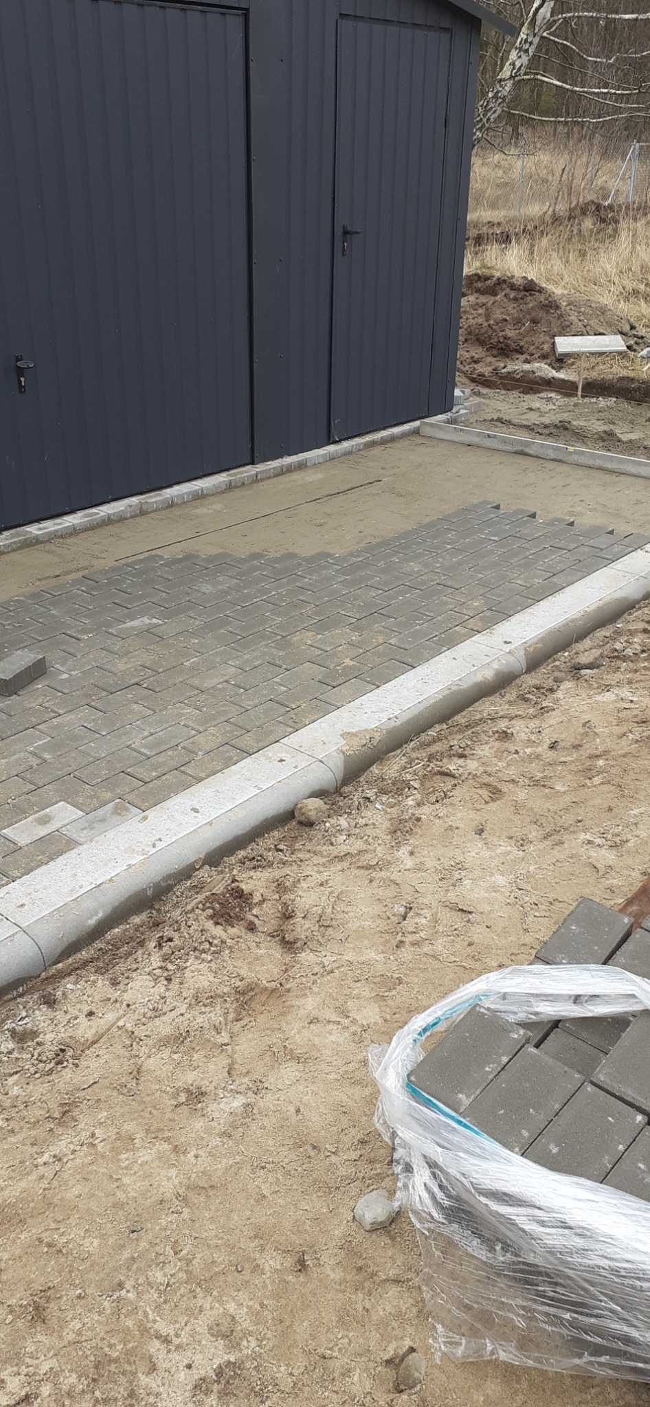 Fragment układanej szarej kostki brukowej w jodełkę, obrzeża z jasnych krawężników, w tle blaszany garaż i ziemia.