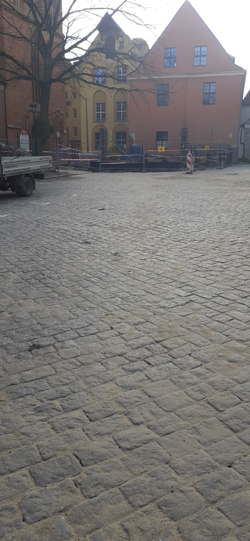 Ułożona kostka granitowa na placu miejskim, ogrodzona barierkami z taśmą ostrzegawczą, w tle budynki starego miasta i zaparkowany samochód dostawczy.
